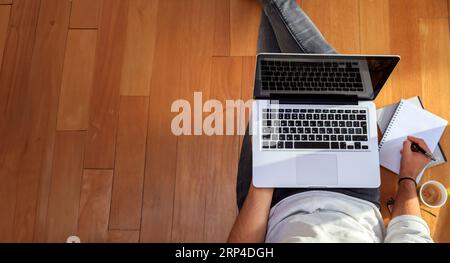 Young man is sitting with black blank screen laptop on wooden floor writing at notebook next to cup of coffee. Workspace at home. Top view, copy space Stock Photo