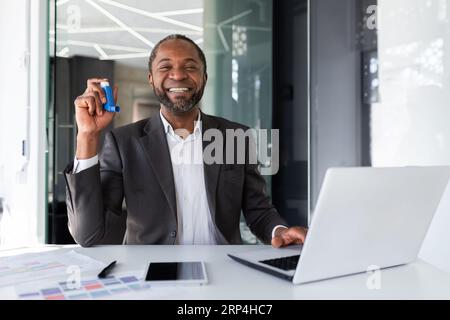 Portrait of satisfied treated man inside office, businessman smiling and looking at camera, holding inhaler for breathing relief and asthma at workplace. Stock Photo