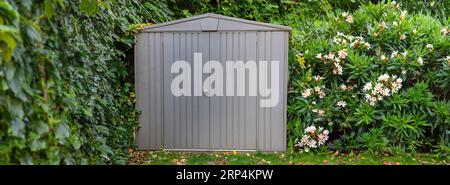 Grey shed, gardening tool storage, gardenhouse at backyard surrounded from green plant. Facade of metal building box in nature. Banner Stock Photo
