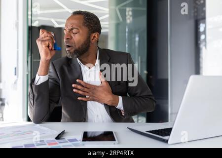 Sick man inside office with asthma using inhaler to ease breathing, mature experienced african american man coughing working sitting with laptop at workplace. Stock Photo