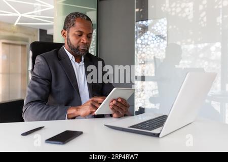 Serious thinking businessman working inside office, using tablet computer, african american mature and confident boss using app at workplace, reading financial performance indicators. Stock Photo