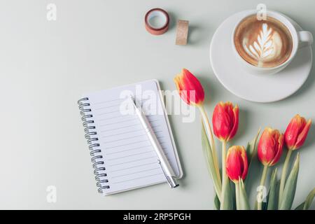 Spiral notepad, pen, coffee cup and red tulip flowers on light green background. Top view, flat lay, mockup. Stock Photo