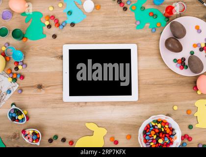 Blank digital tablet surrounded with colorful gems candies easter eggs paper cutout bunny wooden table Stock Photo