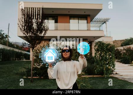 A modern businesswoman in a hijab using VR glasses to check and control her intelligent home, with the futuristic smart house visible in the Stock Photo