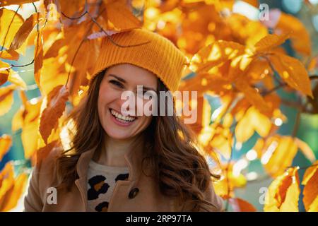 Hello november. happy trendy middle aged woman in brown coat and yellow hat among autumnal foliage outside on the city park in autumn. Stock Photo