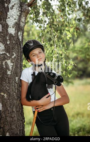 Portrait of a girl - a jockey in a helmet and a white T-shirt, who holds a black horse in her hands - a toy on a stick, looks into the camera. Hobbyho Stock Photo