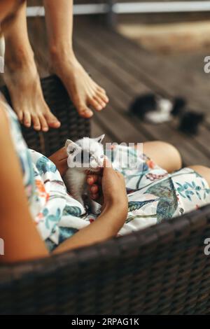 A woman sits in a chair and holds a small kitten in her arms, stroking him. Vertical frame. Stock Photo