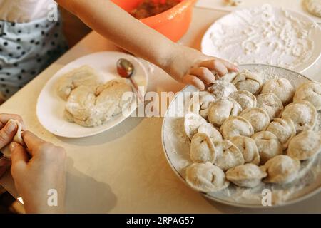 The child puts freshly prepared dumplings on a plate, next to it is dough and flour on the table. Close-up. Stock Photo