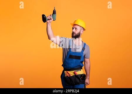 Young carpenter working wtih electric power drill tool, wearing overalls and holding drilling gun. Handyman contractor using cordless drill to work on renovation and refurbishment. Stock Photo