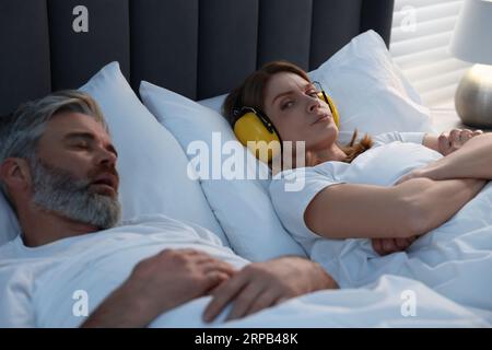 Irritated woman with headphones lying near her snoring husband in bed at home Stock Photo