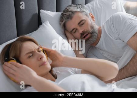 Irritated woman with headphones lying near her snoring husband in bed at home Stock Photo