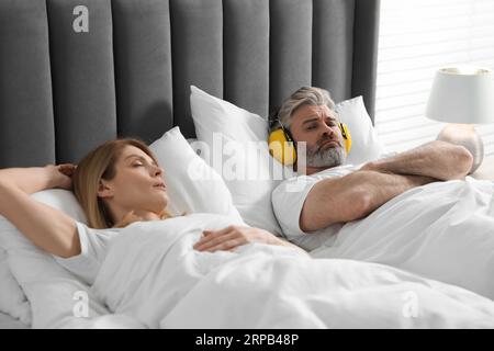 Irritated man with headphones lying near his snoring wife in bed at home Stock Photo