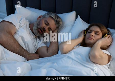 Irritated woman with headphones lying near her snoring husband in bed at home Stock Photo