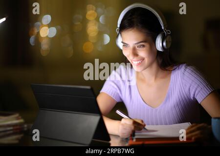 Happy student using tablet and headphone and taking notes in the night at home Stock Photo