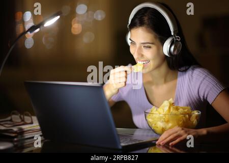 Happy woman in the night wearing headphone eating potato chips watching media on laptop at home Stock Photo