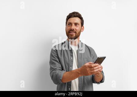 Smiling man looking at side and holding mobile phone while standing on white background Stock Photo