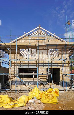 Front elevation new build brick detached house on infill plot takes shape gable wall & roof under construction wrapped in scaffolding Essex England UK Stock Photo