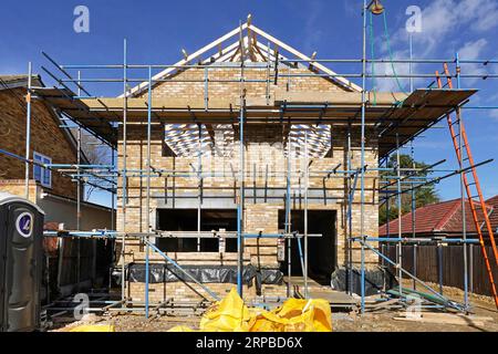 Front elevation new build brick detached house on infill plot takes shape gable wall & roof under construction wrapped in scaffolding Essex England UK Stock Photo