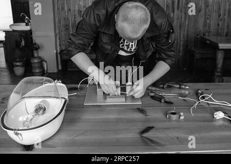 A man working electrician works, collects the electric circuit of a large white street lamp with wires, a relay at an industrial plant factory. Stock Photo