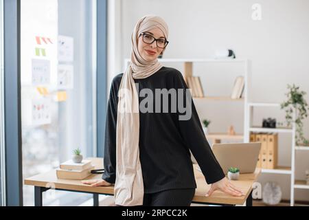 General manager in hijab leaning on desk in spacious office Stock Photo