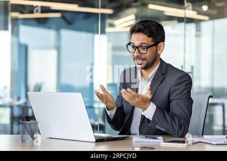 Young successful hispanic businessman talking on video call using laptop, man inside office at workplace, online meeting with colleagues and partners. Stock Photo