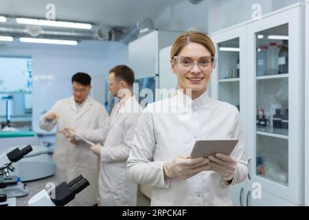 Portrait of a smiling young woman scientist, doctor standing in a clinic laboratory and using a tablet. Behind, male colleagues are discussing work. Stock Photo