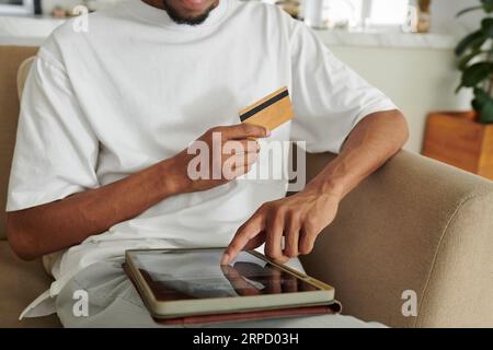 Cropped shot of African American guy with credit card pointing at tablet screen while sitting on couch or armchair and choosing online goods Stock Photo