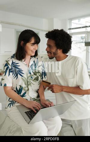 Happy young man with credit card looking at his wife or girlfriend using laptop while consulting with him about chosen goods in online shop Stock Photo