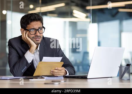 Upset and disappointed with the results of the achievement, young businessman received a mail message, hispanic man desperately reads the news, man works inside the office with a laptop. Stock Photo