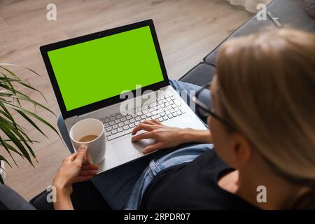 woman sitting on a couch with cup of coffee at home and using laptop computer. blank green screen copy space Stock Photo