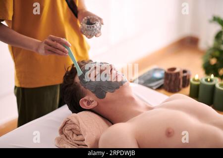 Young female beautician in yellow uniform standing by head of relaxed male client lying on couch and applying clay mask on his face Stock Photo