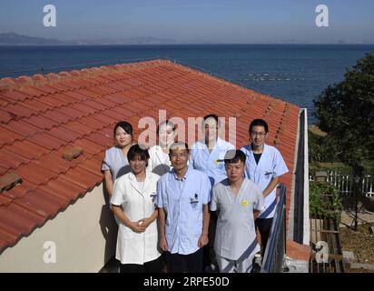 (190818) -- QINGDAO, Aug. 18, 2019 -- Doctor Zhou Zhaojing (C, front) poses for a photo with colleagues in front of the health service center on the Lingshan Island of Qingdao, east China s Shandong Province, Aug. 17, 2019. The Lingshan Island is located some nine sea miles southeast off the west coast of Qingdao. Since 2013, the west coast new area has dispatched medical staff members to work by turns on the island. The 40-year-old Zhou was selected in 2018 as a general practitioner to work at the island s health service center. Despite harsh working and living conditions, Zhou and his collea Stock Photo