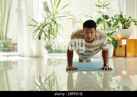 Young active man in striped t-shirt looking at camera while doing push ups on the floor of living room during workout in the morning Stock Photo