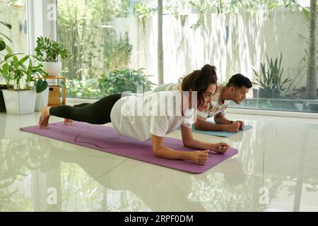 Side view of two young intercultural people doing press ups on mats on the floor of gym or living room with green plants against window Stock Photo