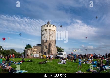 Crowd of spectators watching mass ascent of hot-air balloons at Bristol Balloon Fiesta in sky above Clifton Observatory, Bristol, England Stock Photo