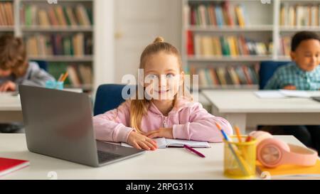 Happy caucasian school girl using laptop at class, looking and smiling at camera, using technology during tech lesson Stock Photo