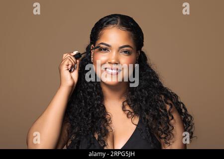Beauty portrait of charming brazilian chubby woman applying mascara on her lashes over brown studio background Stock Photo