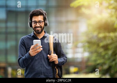 Smiling young indian man wearing headphones holding phone while walking Stock Photo