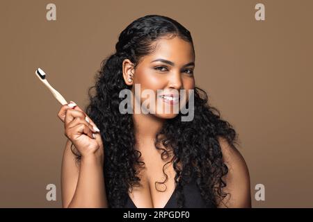 Portrait of beautiful brazilian chubby woman with radiant smile holding wooden toothbrush with paste, brown background Stock Photo