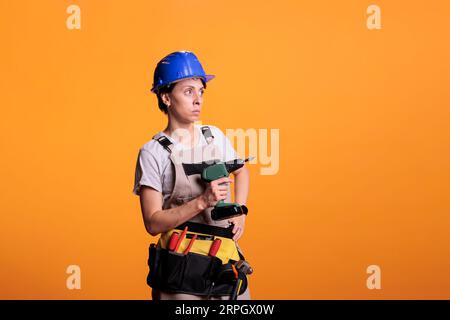 Female construction worker in work uniform with protective helmet holding electric drill on yellow background. Woman professional contractor holding work equipment using tool belt. Stock Photo