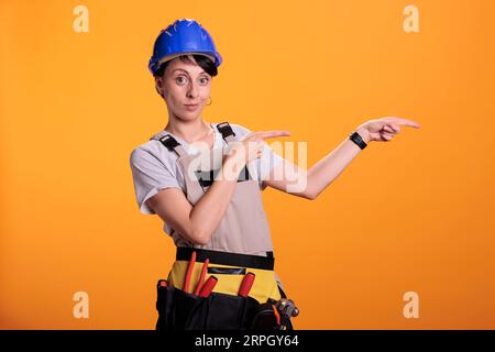 Confident handywoman pointing left or right sides, showing sideways and indicating direction in studio. Builder with helmet posing aside over background, renovation project service. Stock Photo