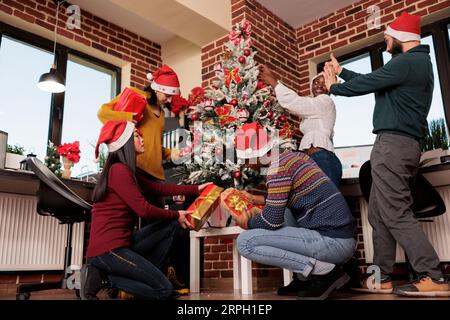 Young startup company employees sharing new year gifts and celebrating holiday in decorated office. Diverse men and women coworkers exchanging presents at corporate christmas party Stock Photo