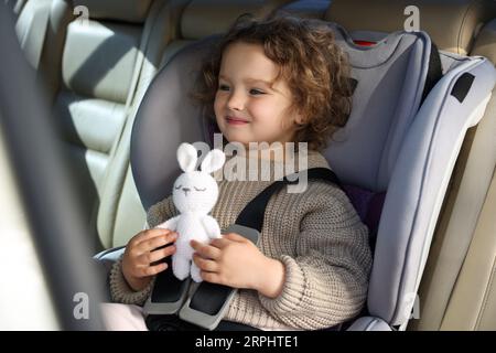 Cute little girl with toy rabbit sitting in child safety seat inside car Stock Photo