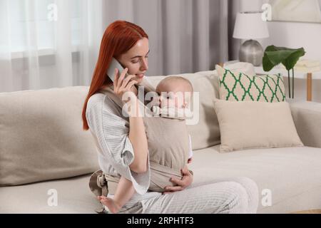 Mother talking on smartphone while holding her child in sling (baby carrier) at home Stock Photo