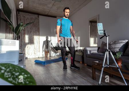 Latin male trainer streaming online workout with phone at home. Online gym class. Stock Photo