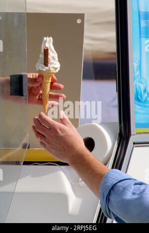 A freshly served 99 Flake is passed down to a happy customer by an ice cream vendor at the van’s serving window during a busy local food festival. Stock Photo