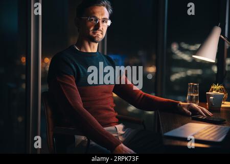 Dedicated businessman looking at the camera while sitting at his office desk after hours. Professional man working overtime to meet his project deadli Stock Photo