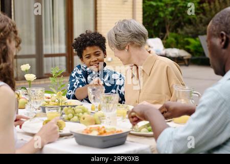 Cute smiling African American boy pointing at empty glass while talking to his grandmother by table served with homemade food and drinks Stock Photo