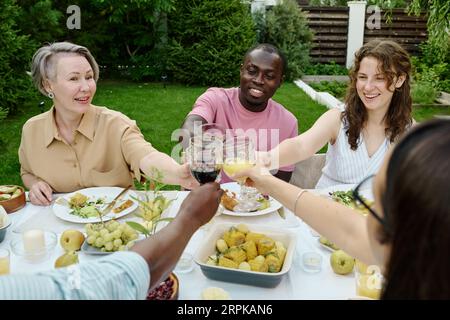 Happy young couple and mature woman sitting by served table in front of their family members and toasting with glasses of red wine with them Stock Photo