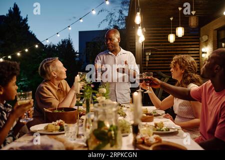 Mature African American man with glass of red wine making toast while standing by served table in front of members of intercultural family Stock Photo